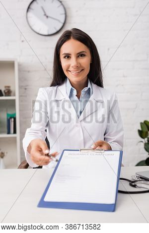 Brunette Doctor Looking At Camera While Holding Pen And Clipboard With Insurance Claim Form