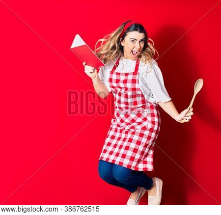 Young beautiful blonde cook woman wearing apron smiling happy. Jumping with smile on face holding recipe book and spoon over isolated red background