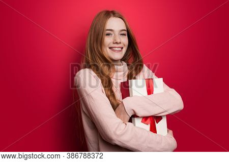 Cute Ginger Woman With Freckles Embracing A Present And Smile At Camera On Red Studio Wall