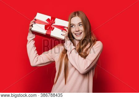 Caucasian Woman With Freckles And Red Hair Is Shaking A Present Posing On Red Studio Wall