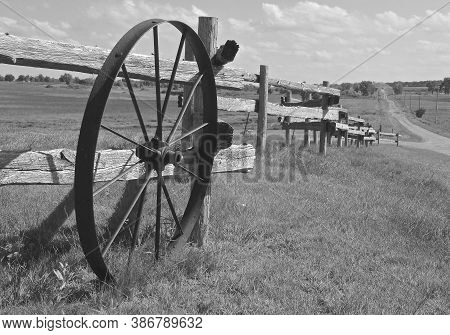 An Old Spoked Wagon Wheel, Rests Along A Weathered Wooden Fenceline Out In The Countryside. (black A