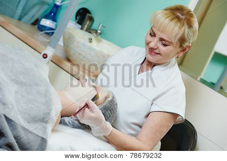 beautician worker with paintbrush applying facial cosmetic mask to female client in beauty salon