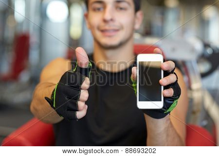 sport, bodybuilding, lifestyle, technology and people concept - happy young man with smartphone showing thumbs up in gym