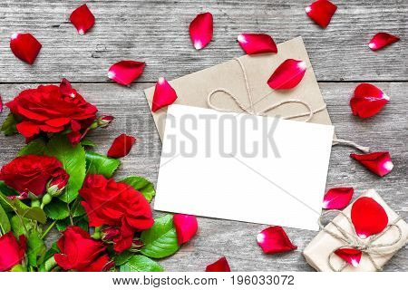 blank white greeting card and envelope with red roses flowers petals and gift box over rustic wooden table. mock up. top view
