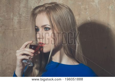 Girl drinking glass of red liqueur. Woman with blond long hair make up in blue dress on beige wall. Alcohol appetizer bad habits addictive and convive. Unhealthy lifestyle