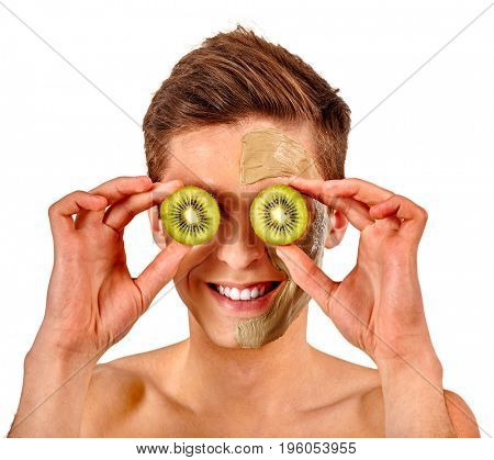 Facial mask from fresh fruits and clay for man concept. Face with treatment mud applied. Male holding kiwi half for skin care procedure in salon. Healing properties of fresh fruit.