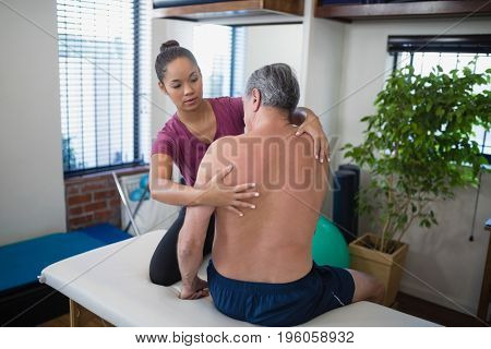 Young female therapist examining back of shirtless senior male patient sitting on bed at hospital ward