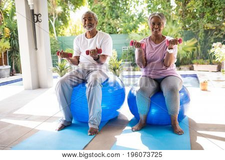 Portrait of senior couple lifting dumbbells while sitting on fitness ball in yard