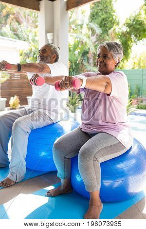 Full length of senior couple lifting dumbbells while sitting on fitness ball in yard