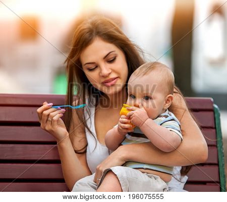 Baby feeding spoon by mother in park outdoor. Weaning in first weeks. Eating child summer on bench. Good appetite by baby food with foster mother number one. Color tone on shiny sunlight background.