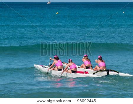 Mount Maunganui, New Zealand - January 28, 2012;  Four women in pink tops paddle surf outrigger canoe through surf in competitions at Mount Maunganui Surf Lifesaving 2012.
