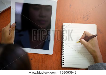 Beautiful Asian businesswoman was looking at the tablet and taking notes on her desk on the wood table in her office. Finance Banking and Technology Concepts.