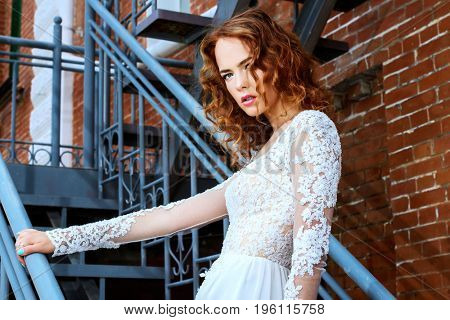 Beautiful young woman with bright foxy hair wearing evening white dress on a city street. Charming bride woman posing outdoor. Urban fashion. 