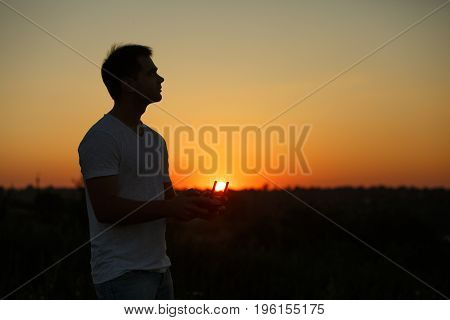 Silhouette of a man piloting drone with a remote controller in his hands on sunset. Pilot takes aerial photos and videos with quadcopter from above.