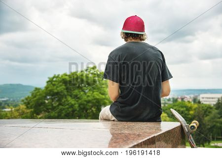 The long-haired skater in a cap and T-shirt sits with his back to the camera and thinks next to the skateboard against the background of the landscape landscape.