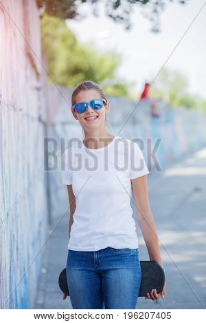 Hipster girl wearing blank white t-shirt, jeans and sunglasses posing against rough street wall, minimalist urban clothing style, mockup for tshirt print store