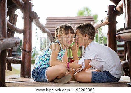 Happy two caucasian siblings sittign in a wooden house outdoors on summer day holding a paper boats in their hands boy making psst gesture