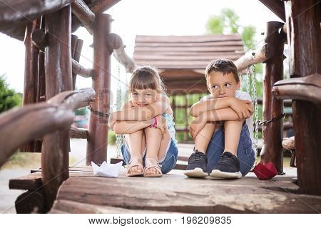 Two young caucasian children sitting next to each other with bent knees in a wooden house outdoors on sunny summer day. They look innocent