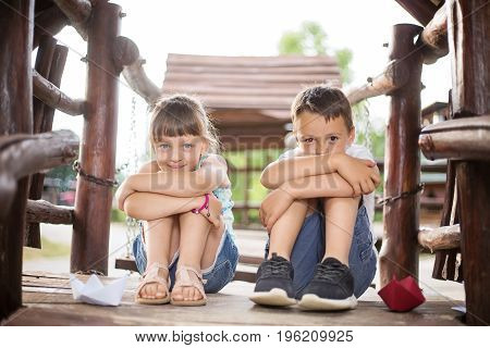 Two young caucasian children sitting next to each other with bent knees in a wooden house outdoors on sunny summer day. They look innocent