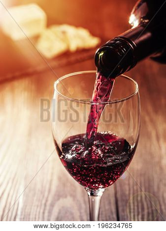 Closeup of red wine pouring in glass on wooden table. heese on background. Copy space. Sunflare effect