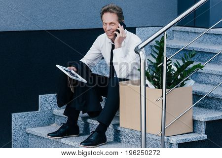 Fired Middle Aged Businessman Holding Photo Frame While Sitting On Stairs And Talking On Smartphone