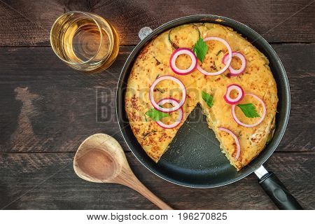 An overhead photo of a Spanish tortilla with potatoes and zucchinis in a tortillera, with a missing slice, with a glass of white wine and a wooden ladle, with a place for text