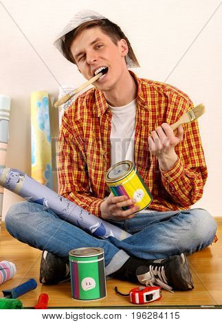 Repair home man holding paint roller for wallpaper. Male in newspaper cap renovation apartment. Paint can and roll paper around. Student loves to work at construction site.