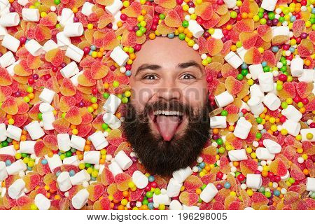 Bearded funny man all covered with sweets and marshmallow putting out tongue and looking at camera.