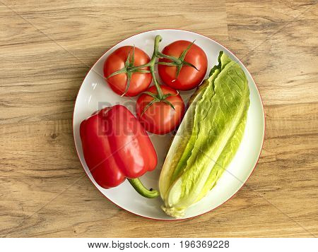 Whole fruits of fresh vegetables close-up: Bulgarian pepper tomato branch and romaine salad lie on white round porcelain plate on the background of a wooden table