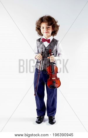 Cute curly violinist in waistcoat holding a violin and looking at the camera. Full height. Gray background.