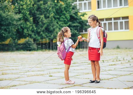 The older sister shares her apple. Two girls in the background of the school.