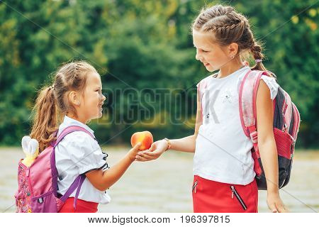 The older sister shares her apple. Two girls in the background of the school.