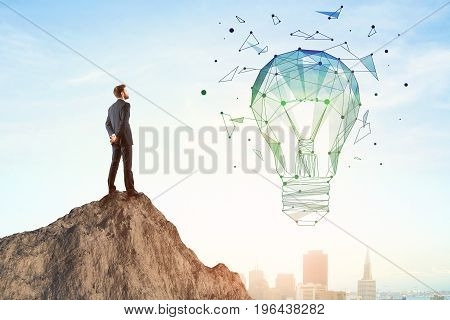 Back view of businessman on mountain top looking at abstract polygonal lamp. City and bright sky background. Innovation and idea concept