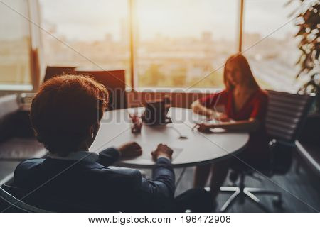 Office scene: company female employee is giving a report on work of her team to male boss in formal suit which is sitting with his back to camera in glass meeting room on high floor of skyscraper