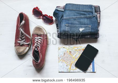 Travel accessories: hipster sneakers, denim shorts, plaid shirt, cowboy belt, hat, sungkasses, map and phone with headphones on wooden background. Selective focus. Traveling concept
