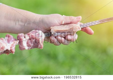 The middle aged woman hand puts pieces of meat for a shish kebab barbeque on a skewer with green blurry background and sun.