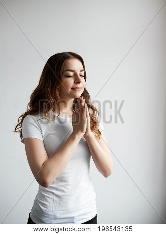 Positive girl wearing white casual t-shirt holding hands in namaste or prayer smiling and keeping eyes closed while meditating and practising yoga indoors having calm expression on her pretty face