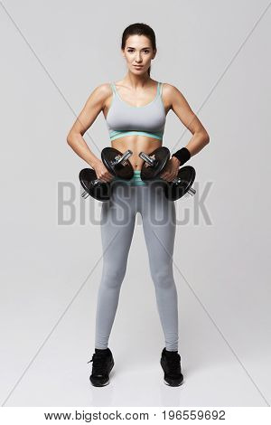 Beautiful sportive girl in sportswear looking at camera straigth holding big dumbbells over white background. Copy space.