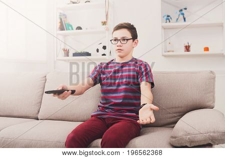 Teenager boy watching television, using remote control to switch channels. Youngster unhappy at what he sees on TV screen. Sitting on couch in living room at home