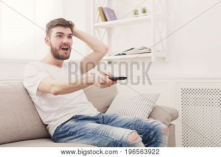 Young man wathing tv. Worried guy pointing with remote controller on tv-set. Man cheering on a favourite football team at home, copy space