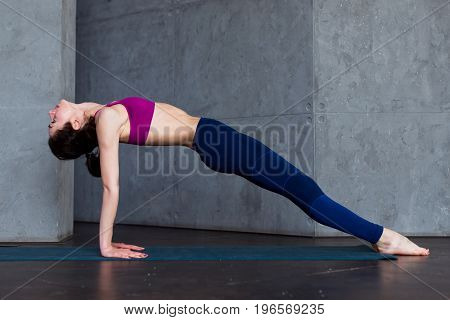 Pretty thin woman in sports bra and leggings doing yoga upward facing plank pose, purvottanasana, working out indoors.