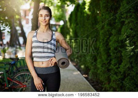 Young female ready to do yoga holding a mat looking at camera smiling. Pilates is good for bodily and mental health.
