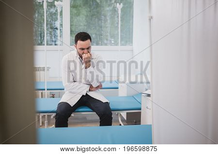 Handsome young doctor in white coat is sitting sadly in hospital ward.