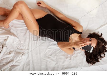 Sexy lazy girl in body underwear lying on the bed taking self photo with mobile phone. Woman relaxing lazing in the bedroom.