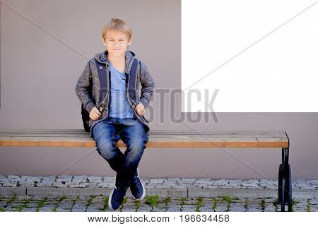 Schoolboy with backpack sitting on the bench near white mock up poster on school wall
