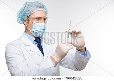 Scrupulous procedure. Capable thorough intelligent man holding up a syringe and preparing for an injection while standing isolated on white background