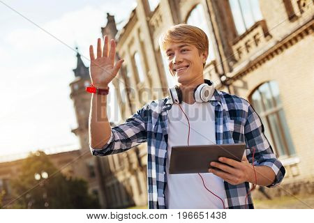 Hello, guys. Friendly active easy going guy spotting fellow students and waving his hand while heading to his lecture with a tablet