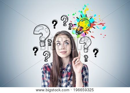 Portrait of a creative young woman wearing a checkered shirt and looking sideways while thinking and pointing upwards. Gray wall background with a bright light bulb and question marks around it.