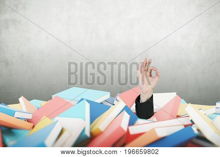 Hand of a man in a suit making on OK sign near a blank concrete wall. He is buried under a pile of book. Mock up