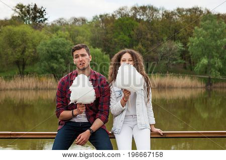 Brunette young man making faces and curly haired woman holding candyfloss sitting at bridge parapet.
